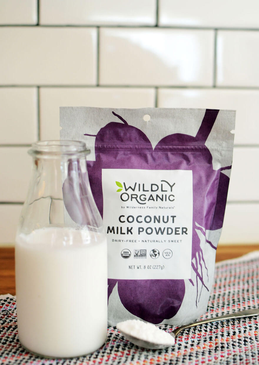 Packaged coconut milk powder with bottle and spoon, against tiled background.