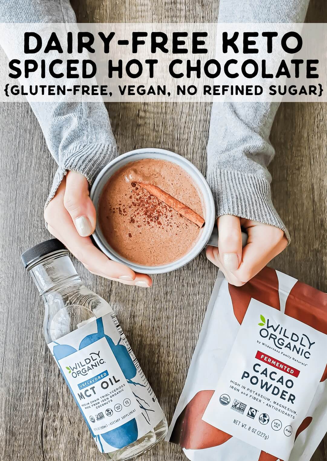 Dairy-Free Keto Spiced Hot Chocolate - Wildly Organic main (1) A photo of two hands holding a mug filled with Dairy-Free Keto Spiced Hot Chocolate with a bottle of MCT oil and a bag of fermented cacao powder.