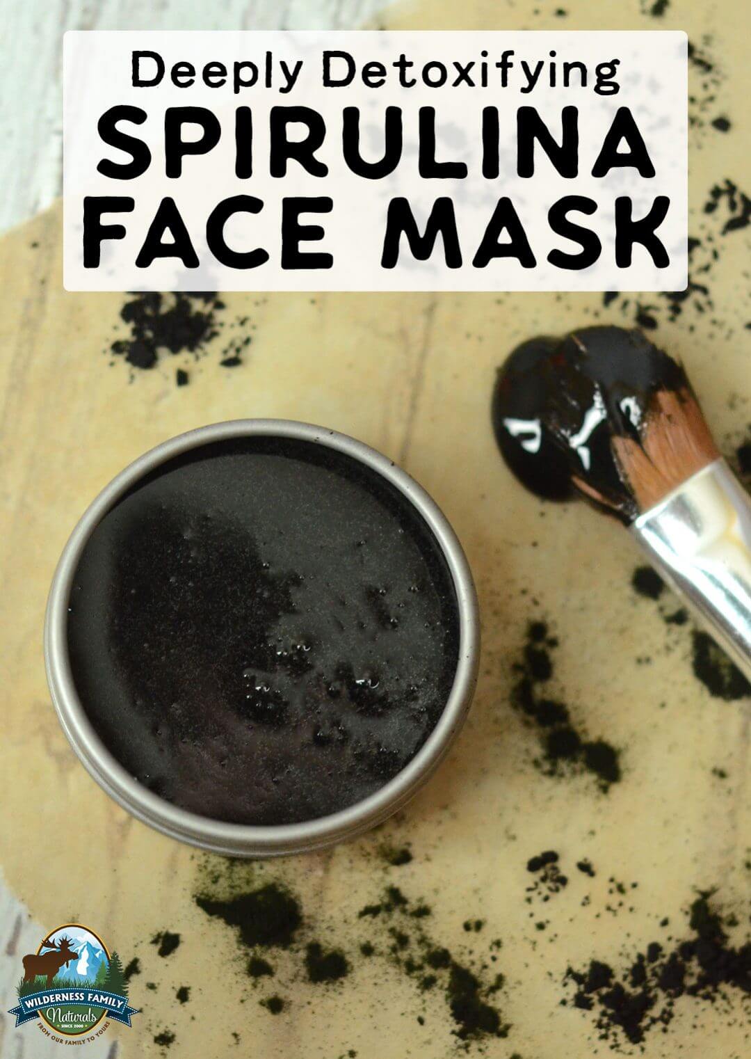 A jar of black spirulina face mask with a brush on a speckled surface and a Deeply Detoxifying label.