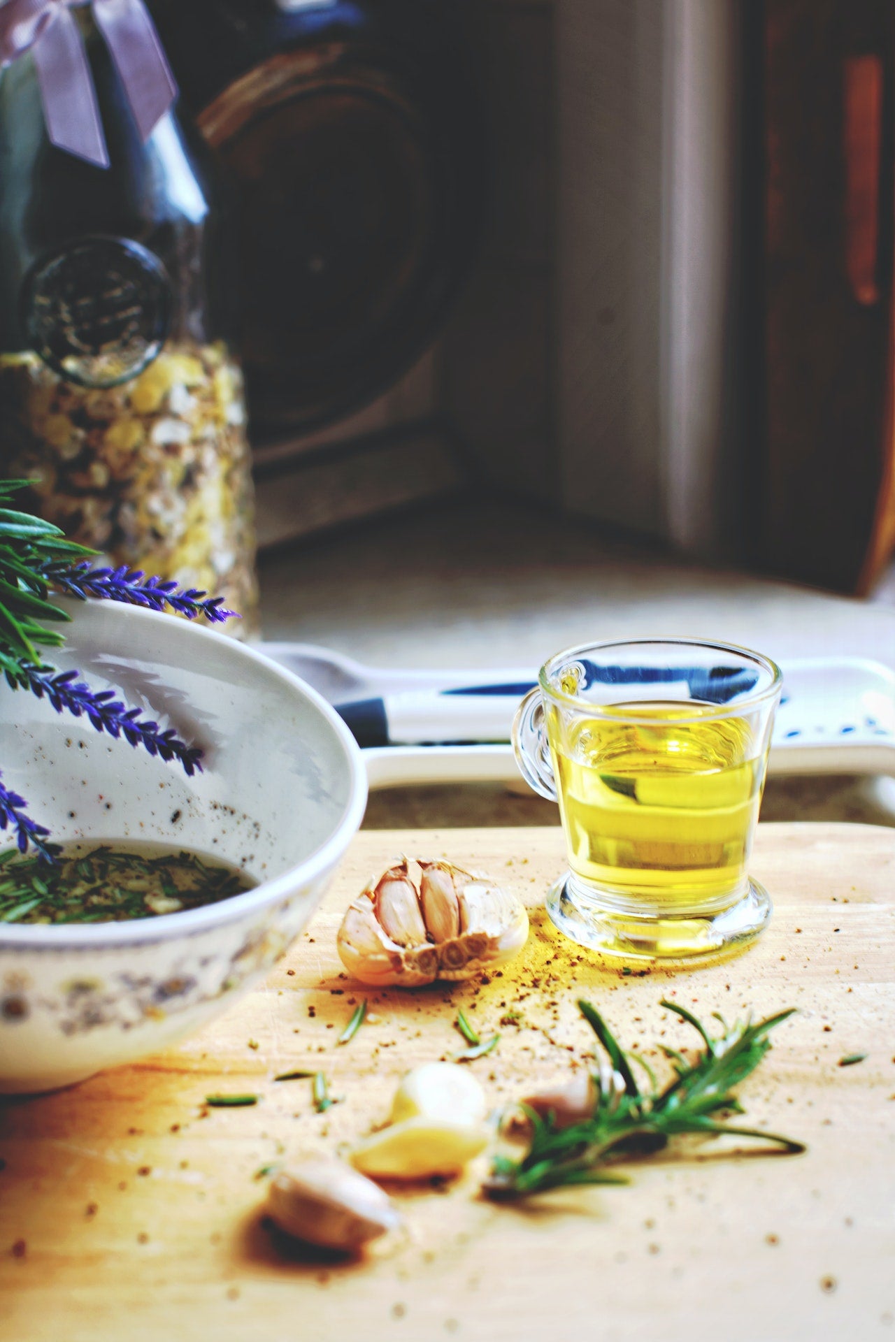 A close-up of a small glass cup of olive oil next to fresh rosemary and garlic cloves, placed on a wooden countertop.