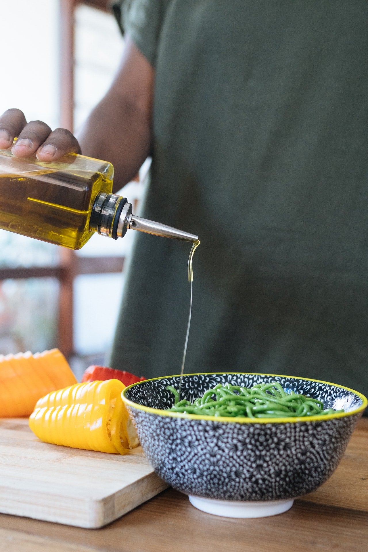 A hand pouring olive oil from a bottle onto green noodles in a bowl, with colorful sliced vegetables in the background.