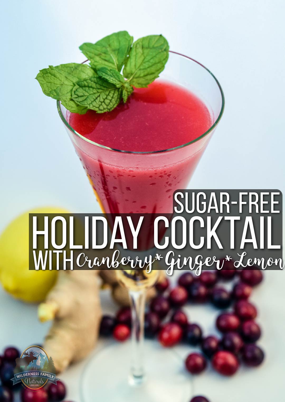 A red holiday cocktail garnished with mint in a martini glass, with cranberries, lemon, and ginger around it