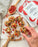 6 of 6:Top down image of Wildly Organic Goji Berry packaging next to goji berry protein balls with chocolate chips and oats on a parchment sheet on a round wooden cutting board with handle and next to a wood spoon and striped dishcloth.