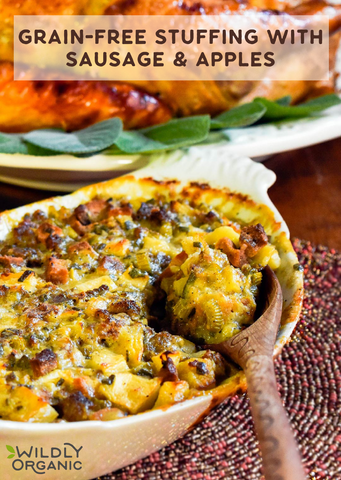 A baked dish of grain-free stuffing with sausage and apples in a white bowl, garnished with sage leaves, and served with a wooden spoon.